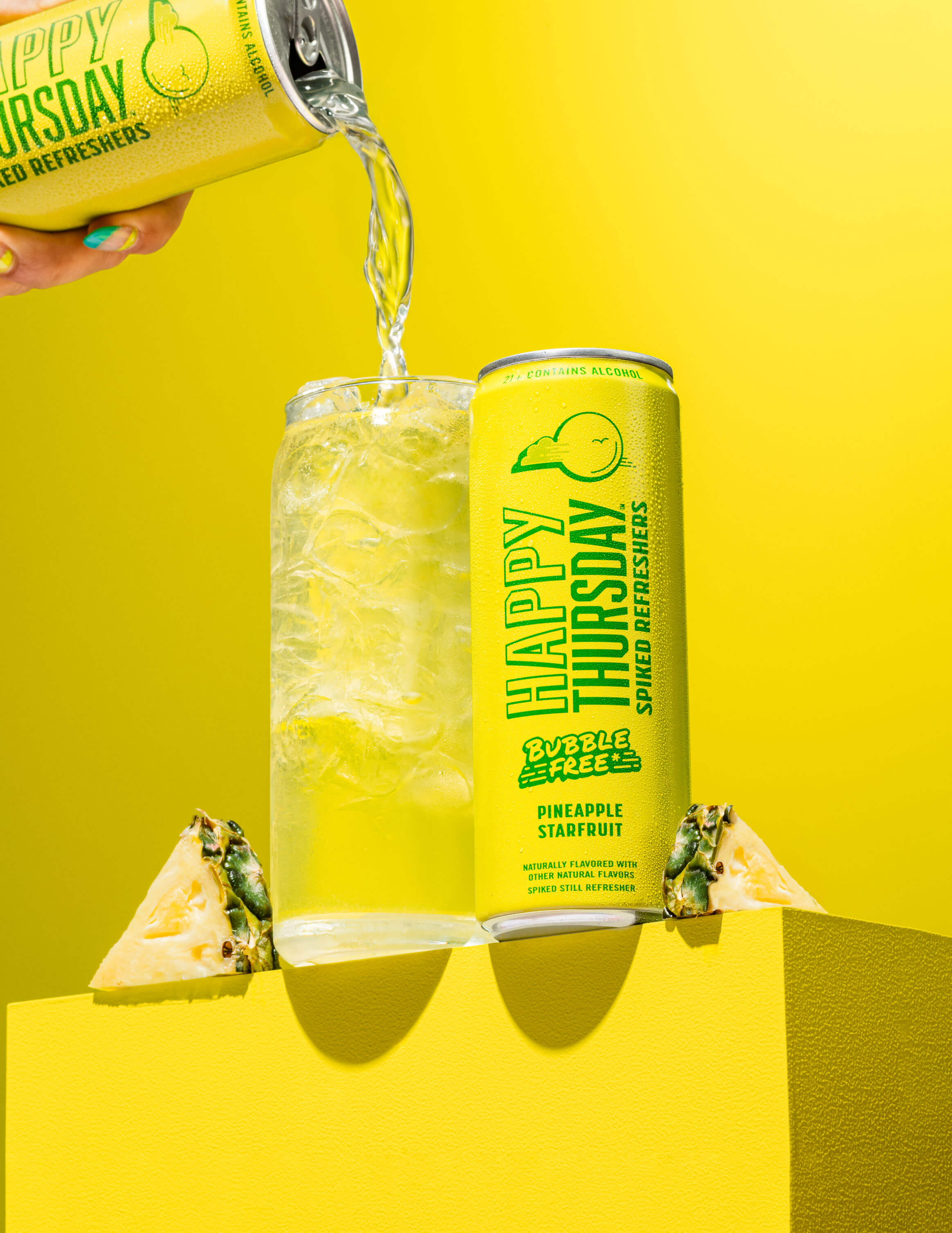 A can of Happy Thursday Pineapple Starfruit is poured over ice into a glass, with Pineapple on a yellow background